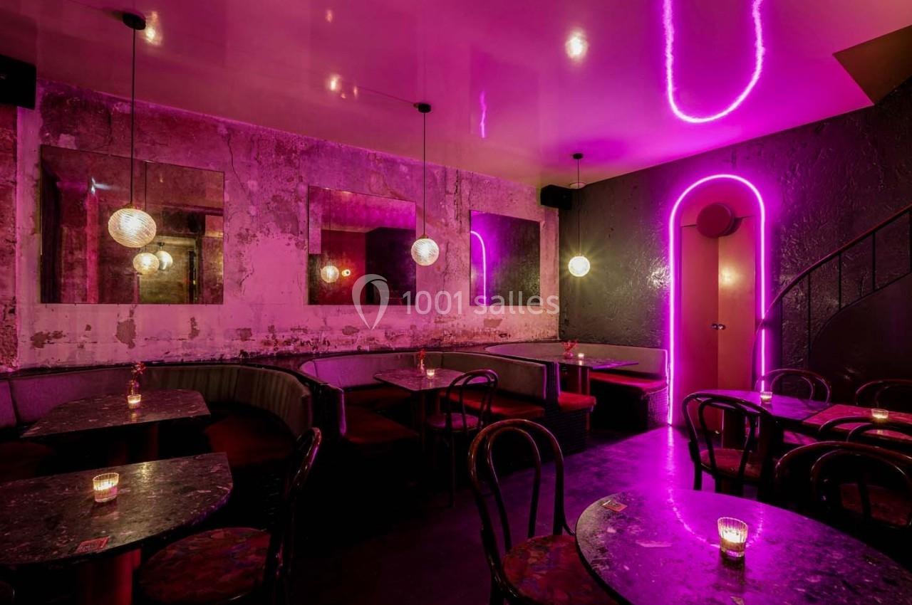Salle de bar avec éclairage néon rose, murs texturés, banquettes, tables en marbre et chaises en bois.