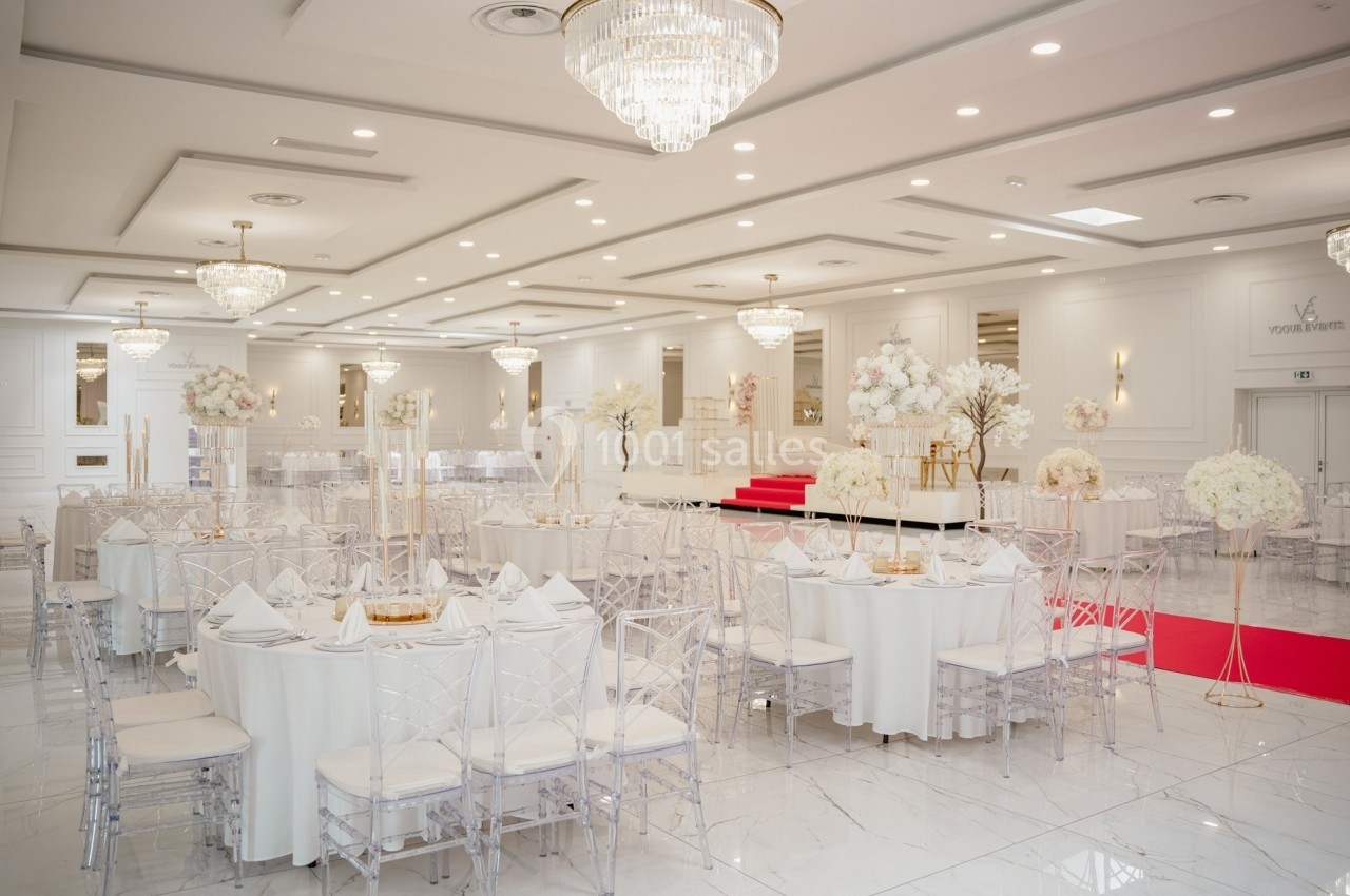 Salle de réception élégante avec tables rondes dressées, chaises transparentes et décorations florales blanches.