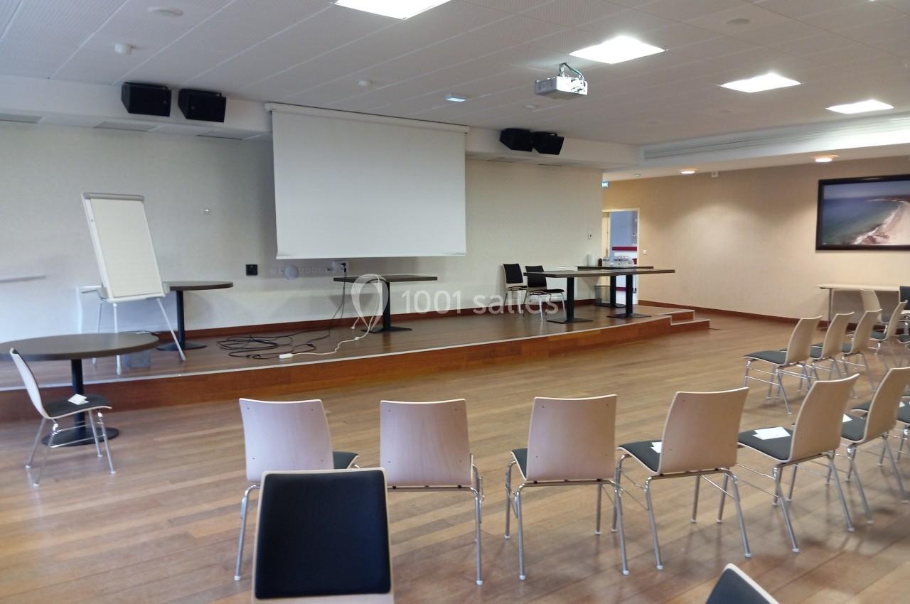 Salle de conférence vide avec chaises alignées, estrade équipée d'un écran de projection, tableau blanc et tables.