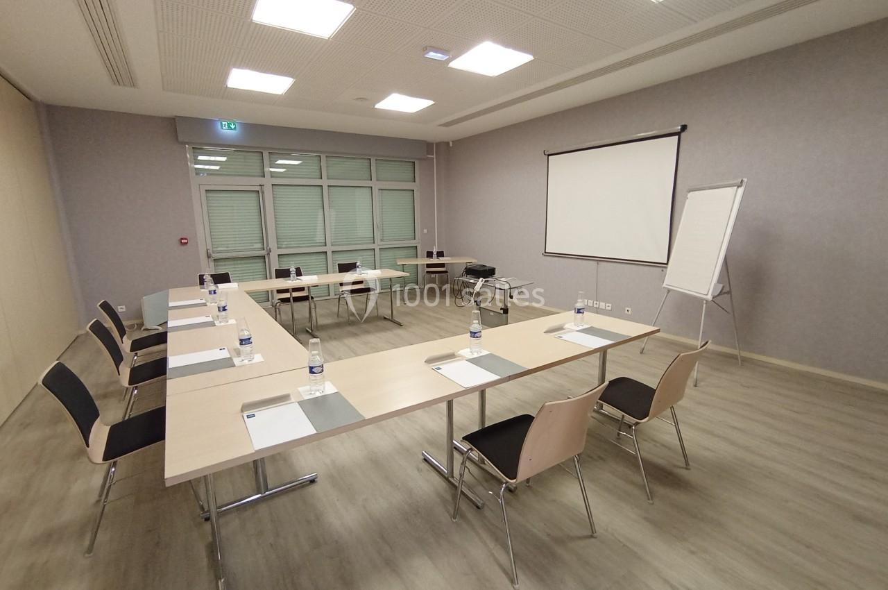 Salle de réunion lumineuse avec tables en U, chaises, écran de projection, tableau blanc et fournitures de bureau.
