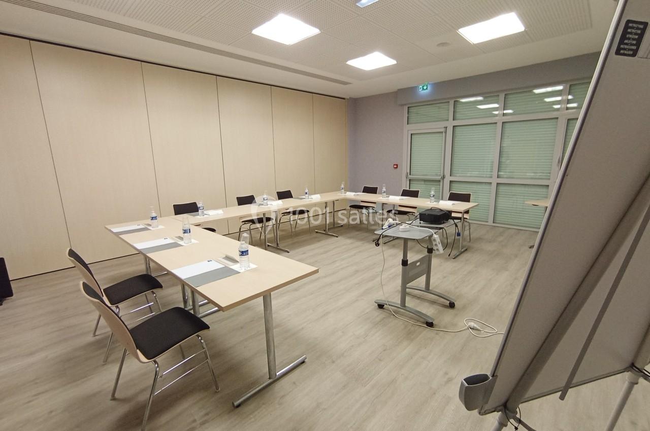 Salle de réunion avec tables disposées en U, chaises, bouteilles d'eau, vidéoprojecteur et tableau blanc.