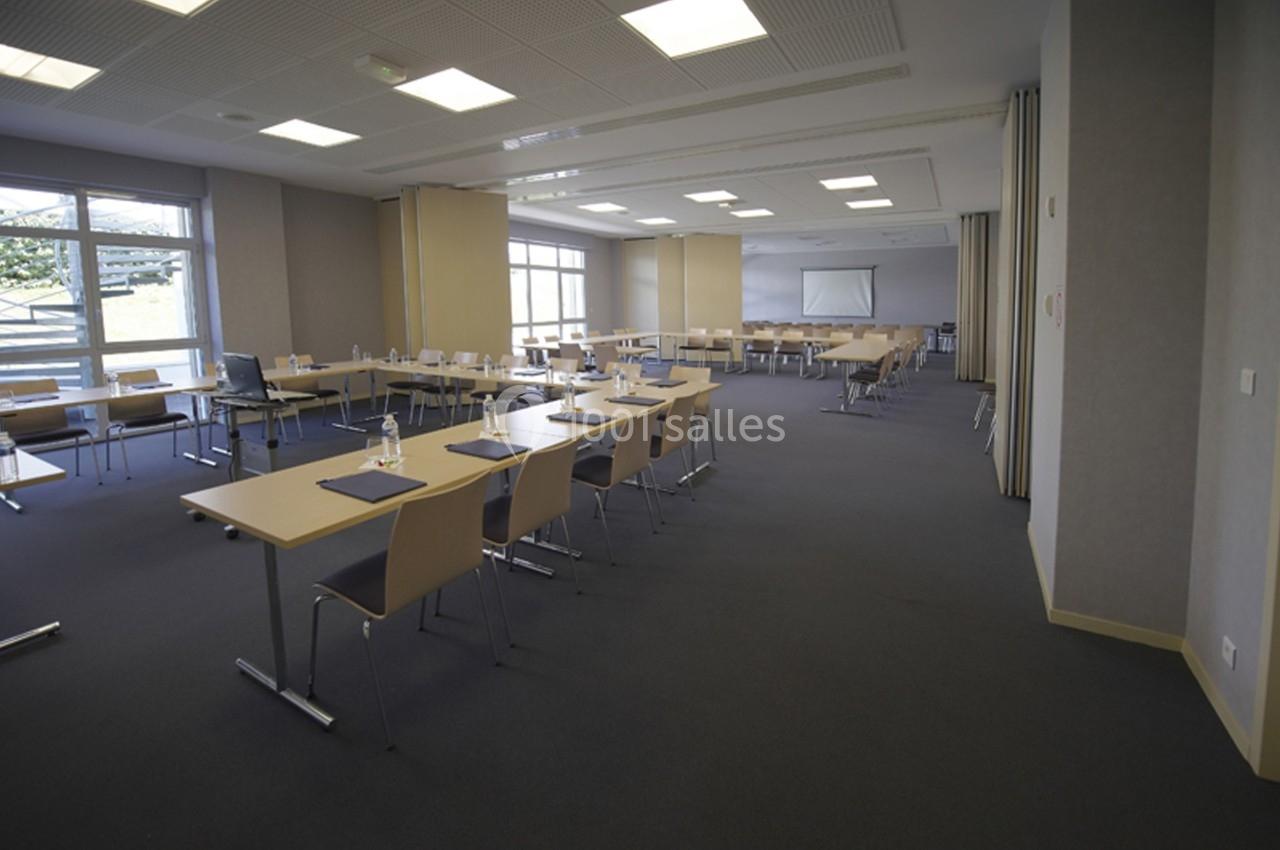 Salle de réunion lumineuse avec des tables alignées, des chaises en bois clair et un écran de projection au fond.