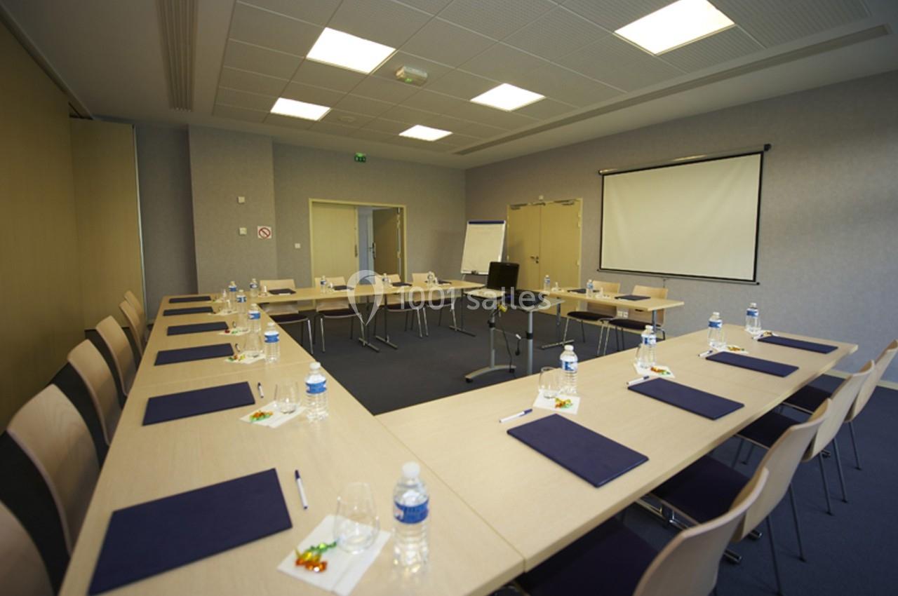 Salle de réunion vide avec tables disposées en U, chaises, bloc-notes, bouteilles d'eau et écran de projection.