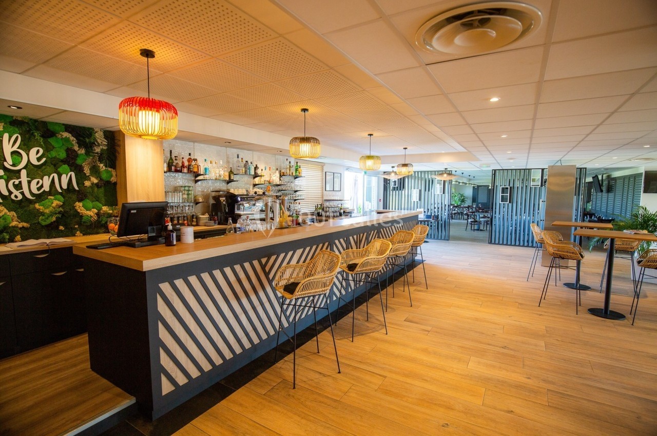 Bar moderne avec comptoir en bois, tabourets en rotin, décor végétal et éclairage suspendu dans une salle lumineuse.