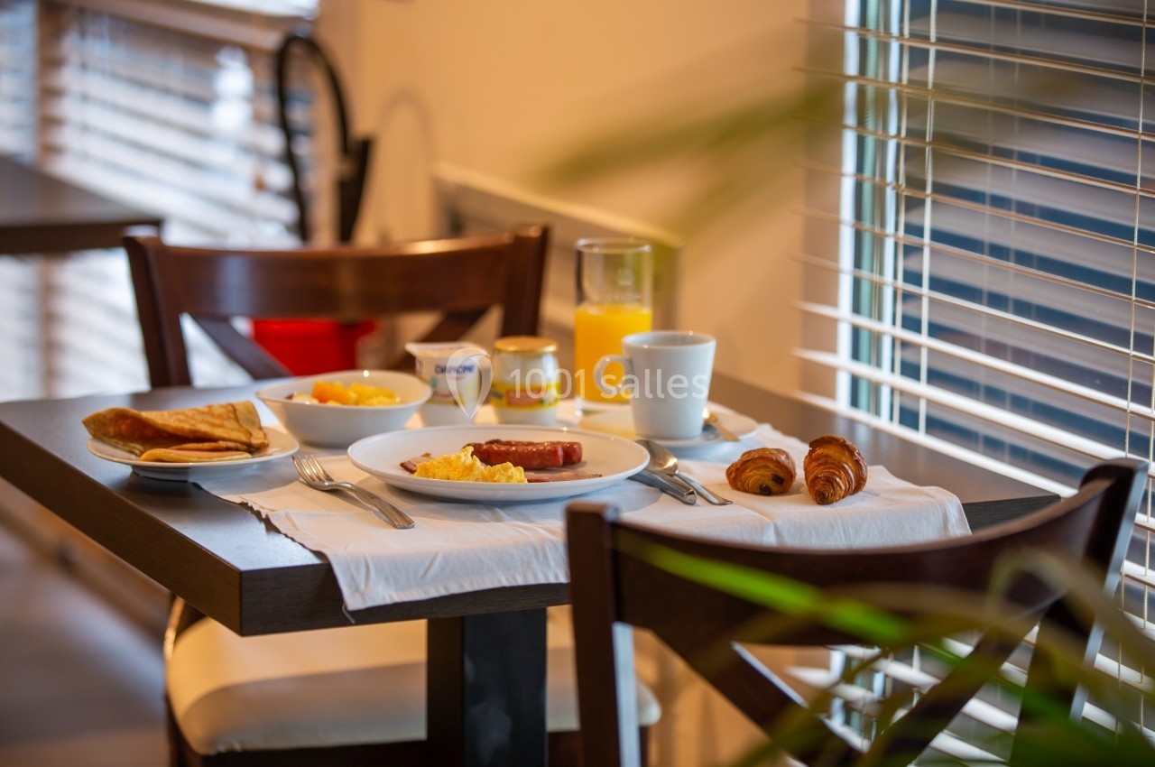 Petit-déjeuner servi sur une table avec croissants, crêpes, œufs, saucisse, jus d'orange et café près d'une fenêtre.
