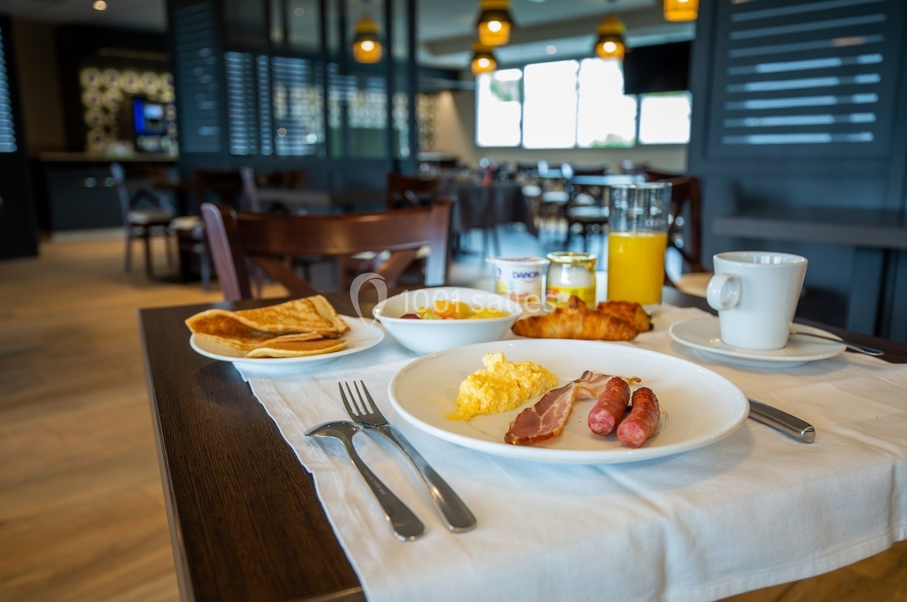 Petit-déjeuner servi sur une table avec œufs, saucisses, croissant, jus d'orange, café et pancakes dans une salle lumineuse.