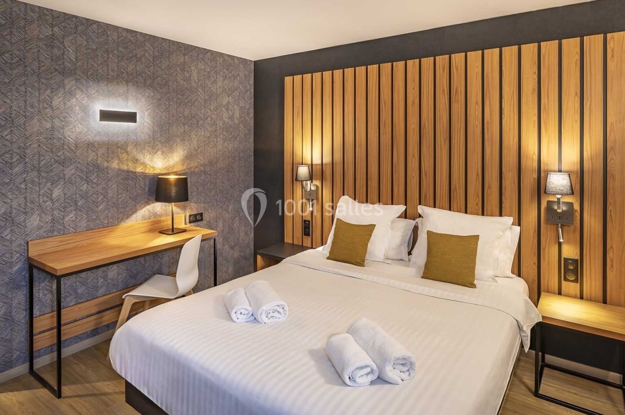 Lit double avec draps blancs, coussins jaunes, deux serviettes pliées, bureau en bois et éclairage mural dans une chambre…