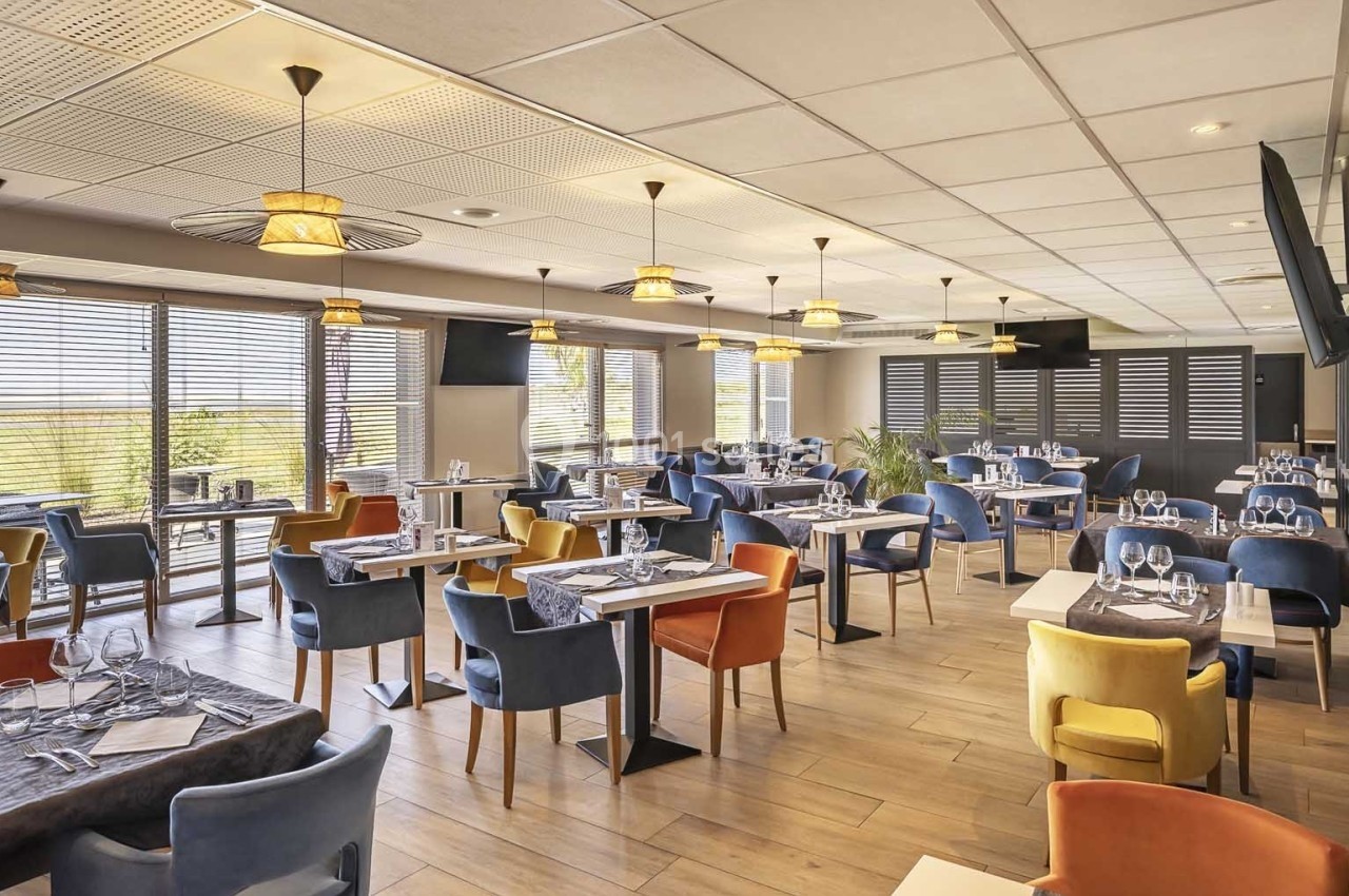 Salle de restaurant lumineuse avec tables dressées, chaises colorées et grandes fenêtres laissant entrer la lumière…