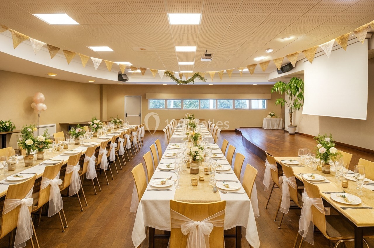 Salle décorée pour un événement, avec longues tables dressées, chaises ornées de nœuds et guirlandes suspendues.