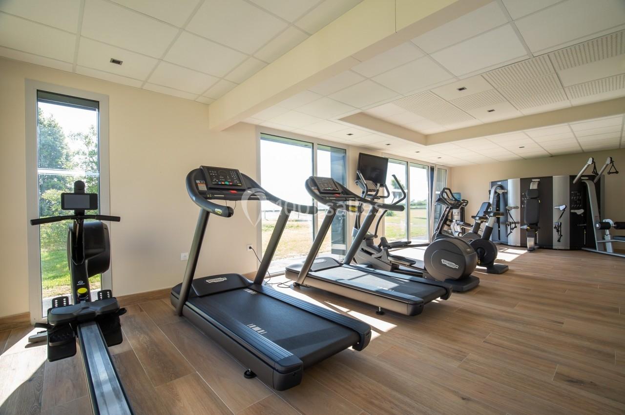 Salle de sport lumineuse avec tapis de course, rameur, vélos d'exercice et équipements de musculation.