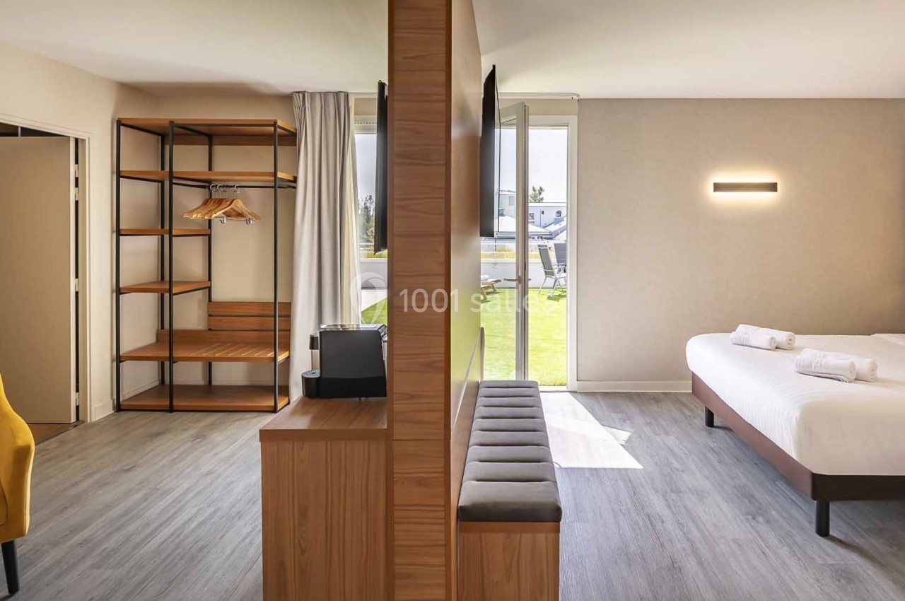 Chambre d'hôtel lumineuse avec lit double, coin salon, étagères ouvertes et vue sur un espace extérieur verdoyant.