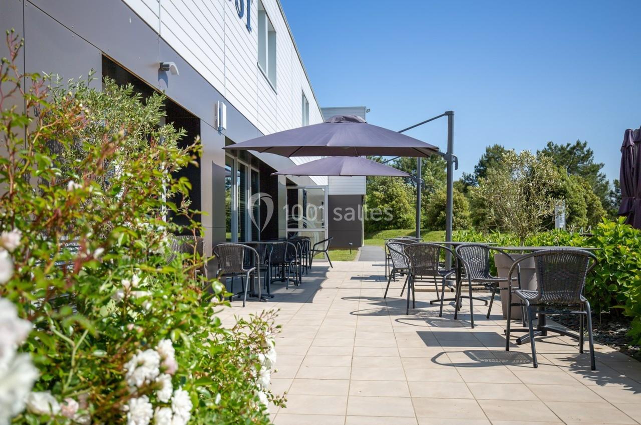 Terrasse extérieure avec tables, chaises et parasols, entourée de végétation sous un ciel bleu.