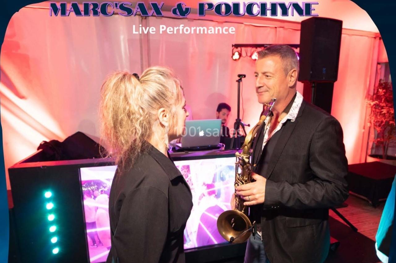 Un homme tenant un saxophone échange avec une femme devant une scène avec un DJ en arrière-plan.