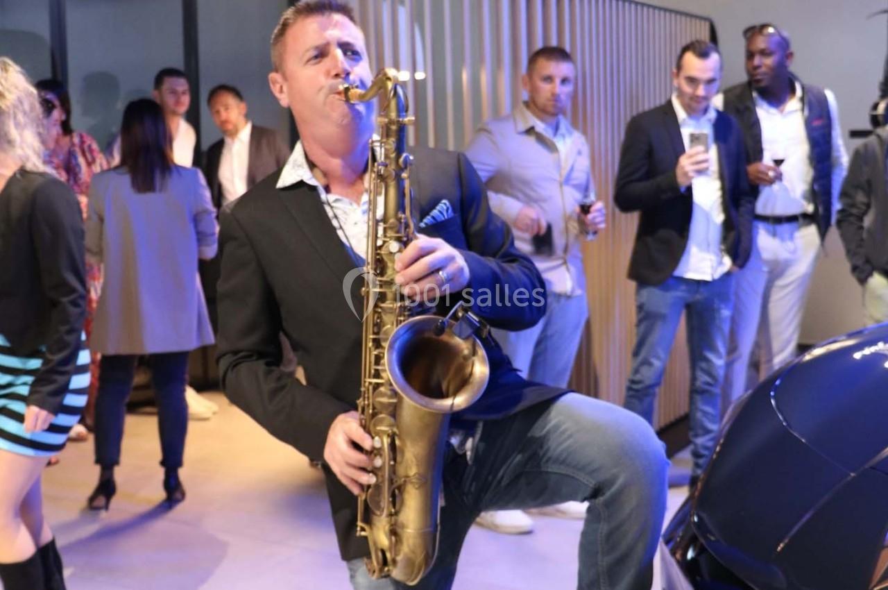 Un musicien joue du saxophone lors d'un événement, entouré de personnes debout dans un espace intérieur moderne.