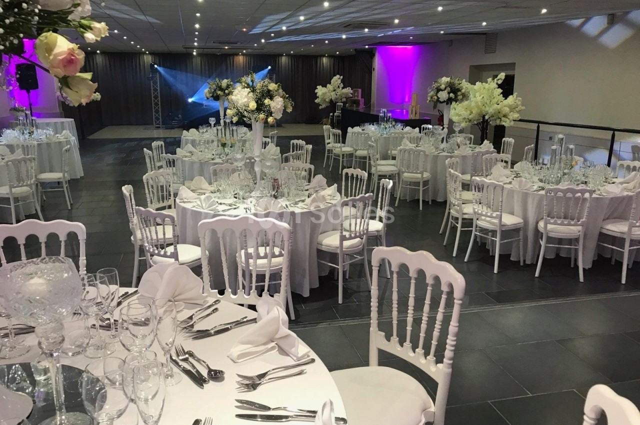 Salle de réception élégamment décorée avec des tables rondes, nappes blanches, chaises assorties et éclairage coloré.