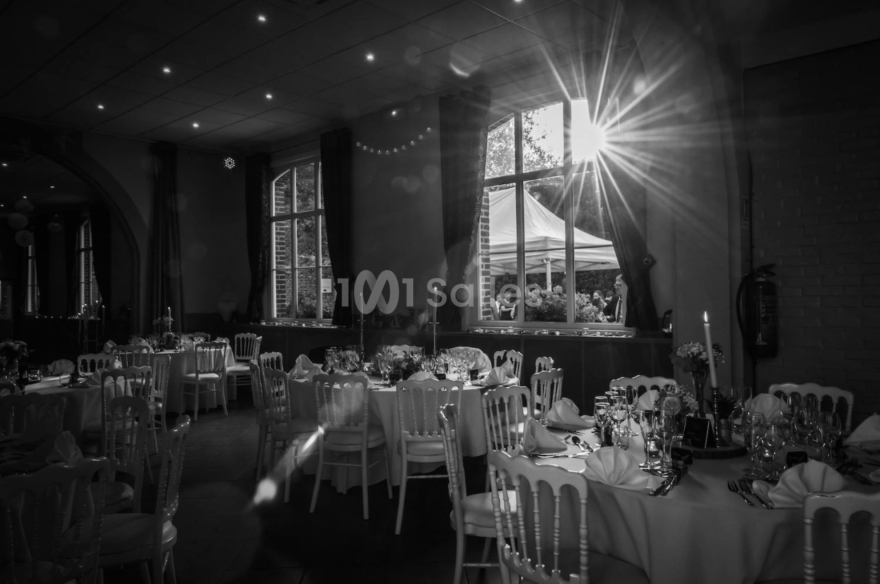 Salle de réception décorée avec des tables dressées, éclairée par la lumière du soleil traversant une grande fenêtre.