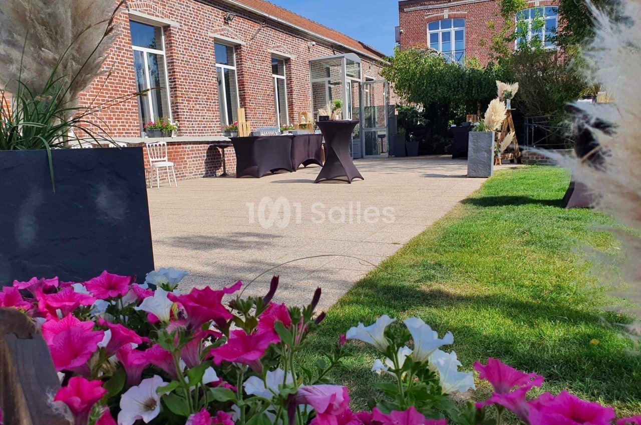 Terrasse ensoleillée avec tables hautes, fleurs colorées au premier plan et bâtiment en briques rouges en arrière-plan.