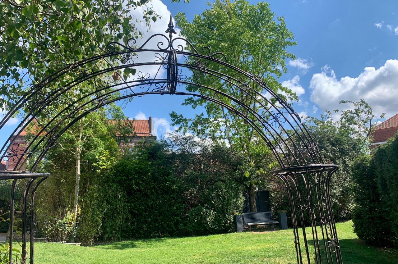 Location salle Comines (Nord) - Les Salons du Château - France Evénement #35 Arche en fer forgé dans un jardin verdoyant avec pelouse, arbres et ciel bleu parsemé de nuages.
