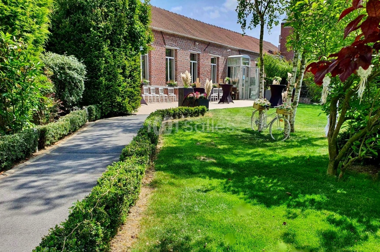 Jardin verdoyant avec allée bordée de haies, donnant sur une maison en briques et une terrasse aménagée.
