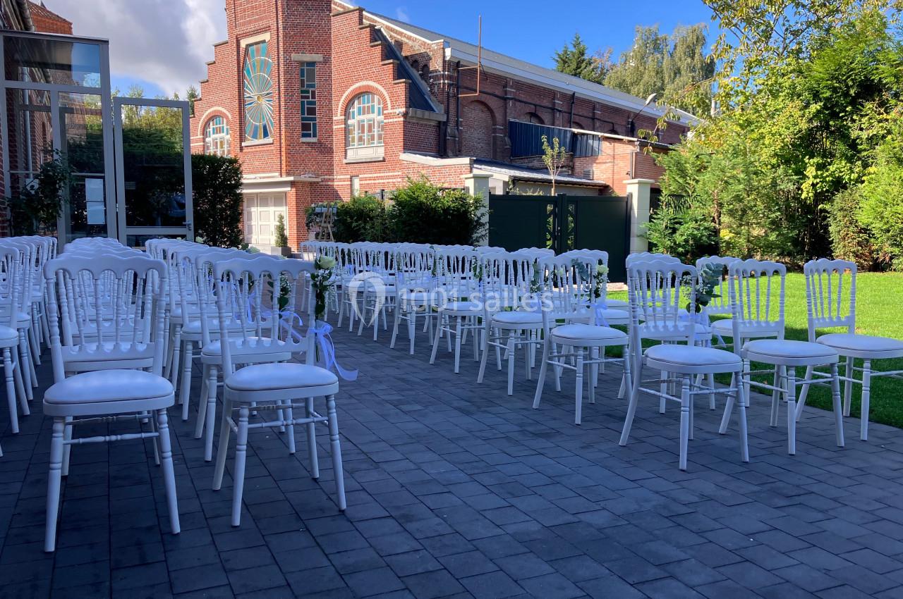 Location salle Comines (Nord) - Les Salons du Château - France Evénement #18 Chaises blanches alignées en extérieur sur une terrasse pavée, avec un bâtiment en briques et un jardin en arrière-plan.