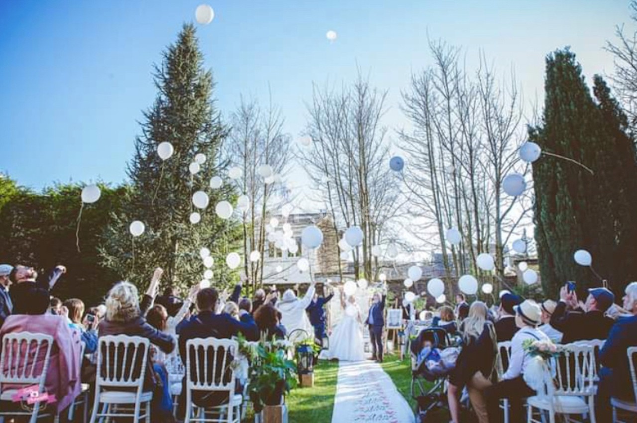 Cérémonie de mariage en plein air avec des invités assis, des ballons blancs lâchés et des arbres en arrière-plan.