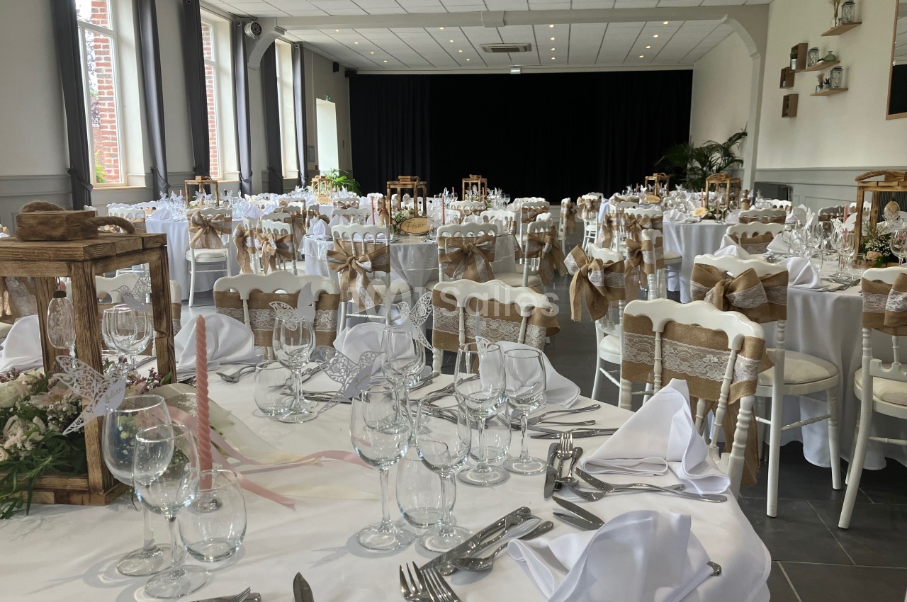 Salle de réception décorée pour un mariage, avec tables dressées, nappes blanches et chaises ornées de rubans beiges.