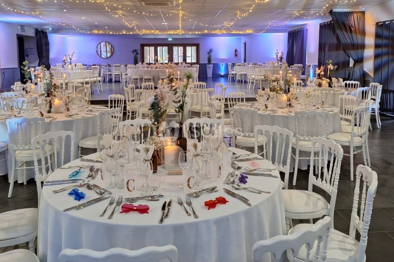 Salle de réception décorée avec des tables rondes dressées, chaises blanches et éclairage chaleureux.