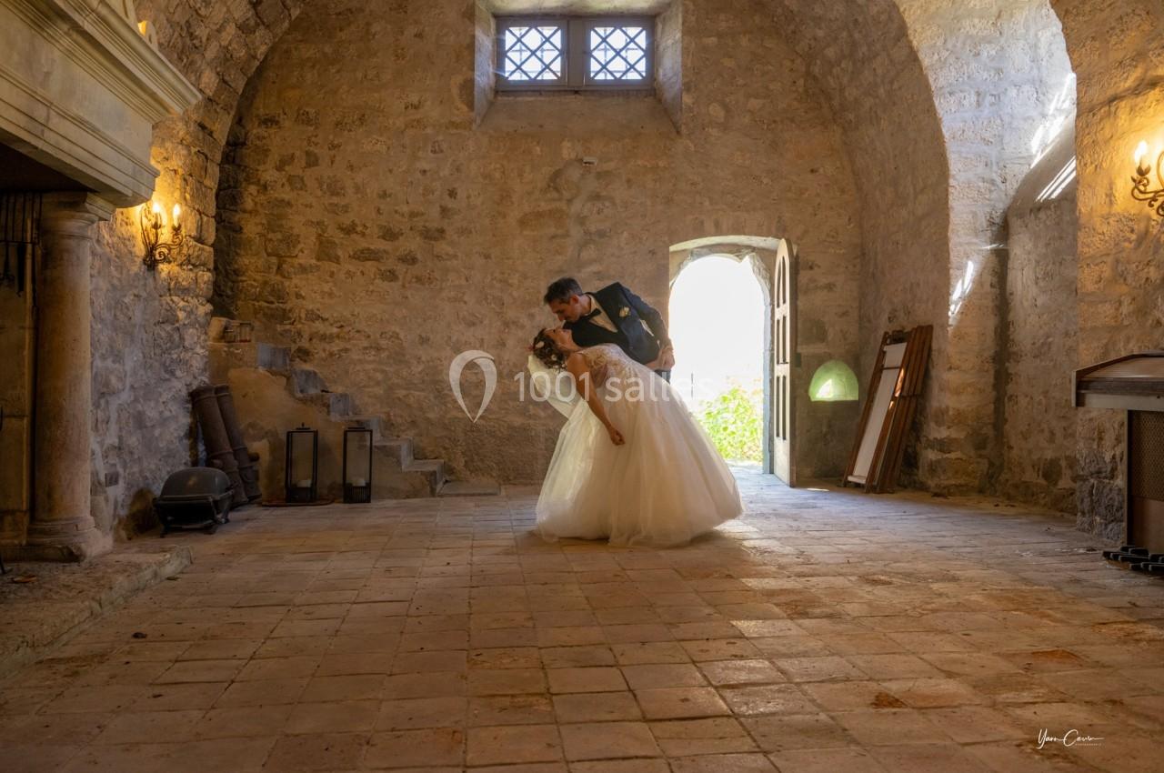 Un couple de mariés danse dans une salle en pierre éclairée par la lumière naturelle entrant par une porte ouverte.