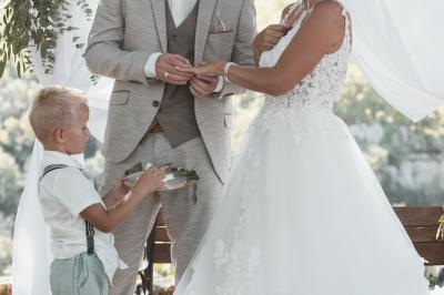 Miniature Corinne & Romain Femme assise, vêtue d'une robe blanche fluide, posant avec une expression sereine sur fond neutre.