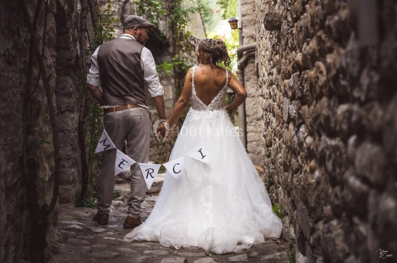 Un couple de mariés de dos, tenant une bannière ’Merci’, dans une ruelle pavée bordée de murs en pierre.