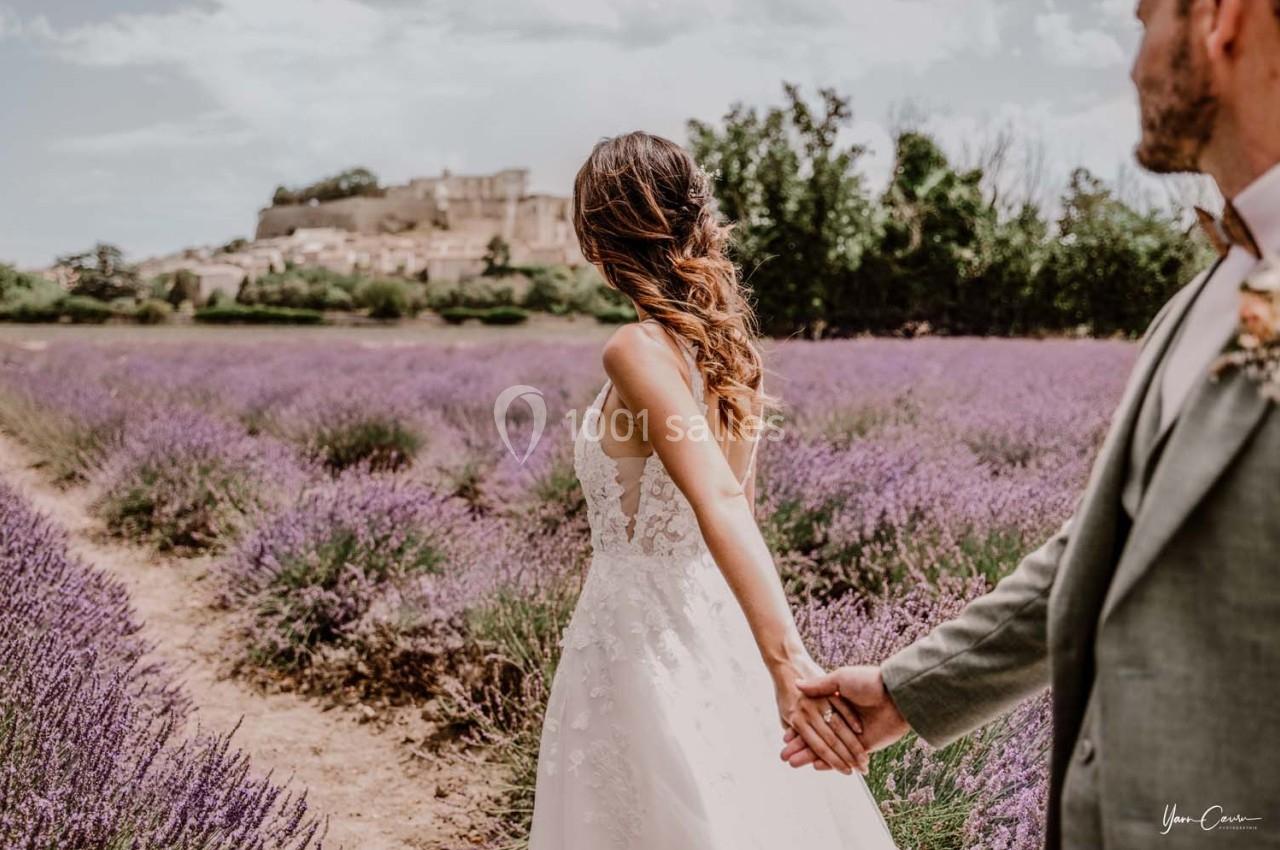 Une femme en robe de mariée tient la main d'un homme dans un champ de lavande avec un village en arrière-plan.