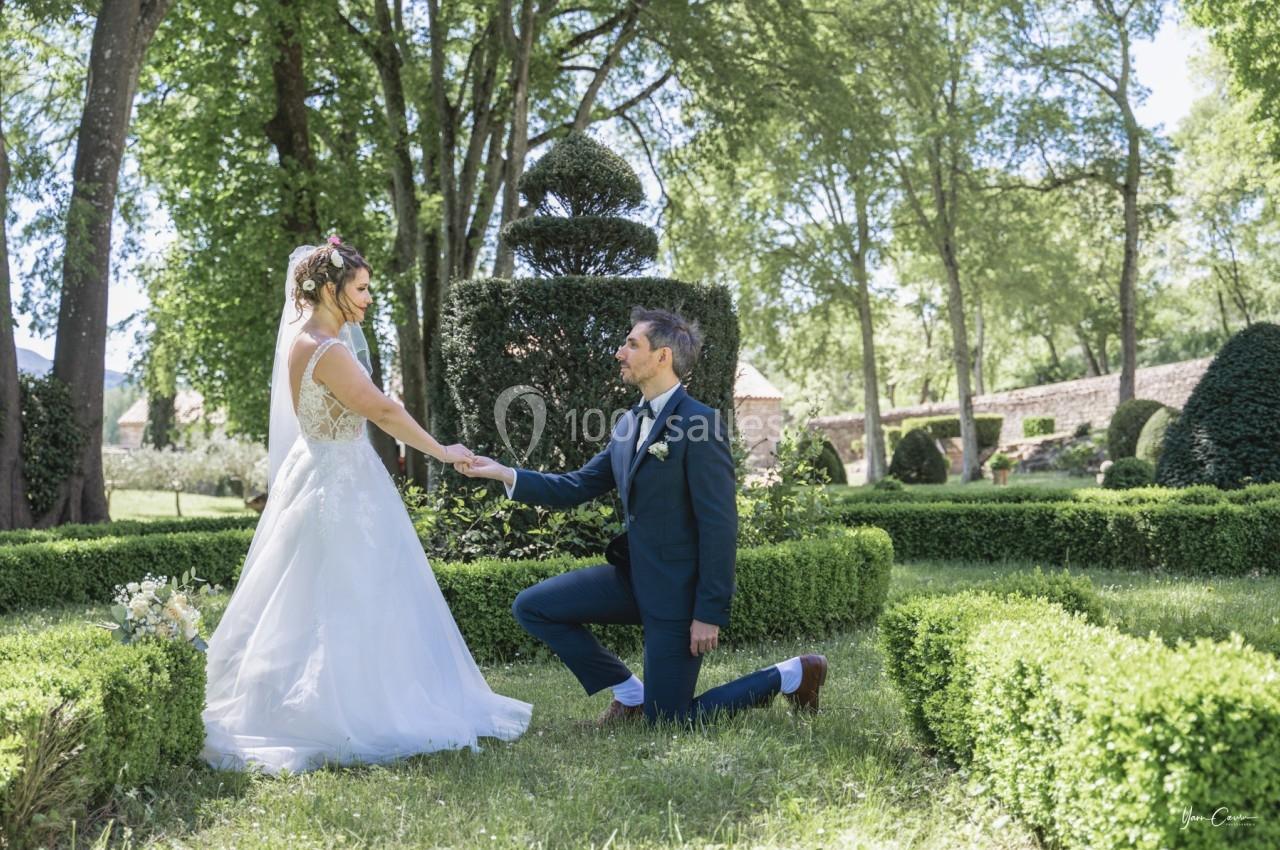 Un marié à genoux tient la main de la mariée dans un jardin avec des haies taillées et des arbres en arrière-plan.