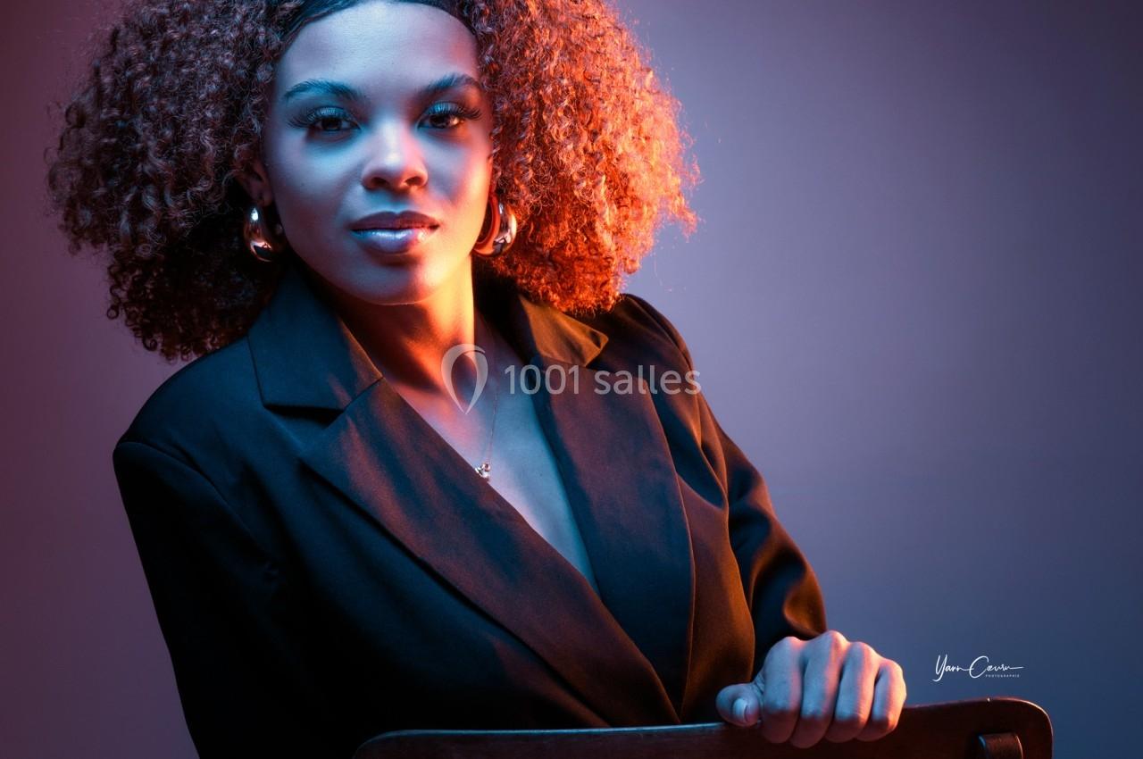Femme aux cheveux bouclés portant un blazer noir, assise sur une chaise, éclairée par une lumière colorée.