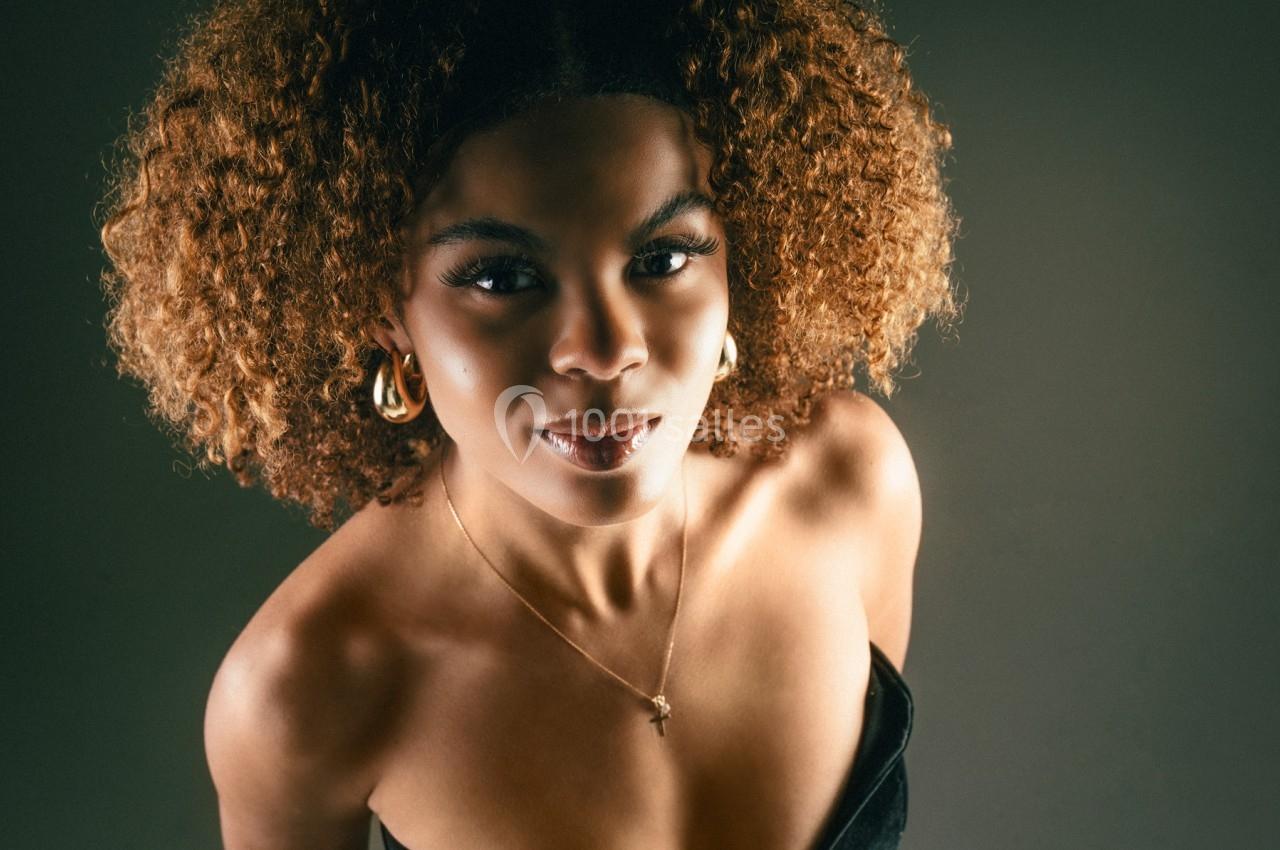 Femme aux cheveux bouclés regardant l'objectif, portant un collier doré et une tenue noire, sur fond sombre.