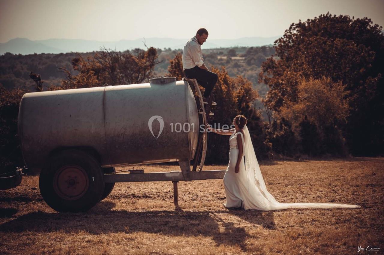 Un couple en tenue de mariage, la mariée touche la main du marié assis sur une citerne dans un champ ensoleillé.