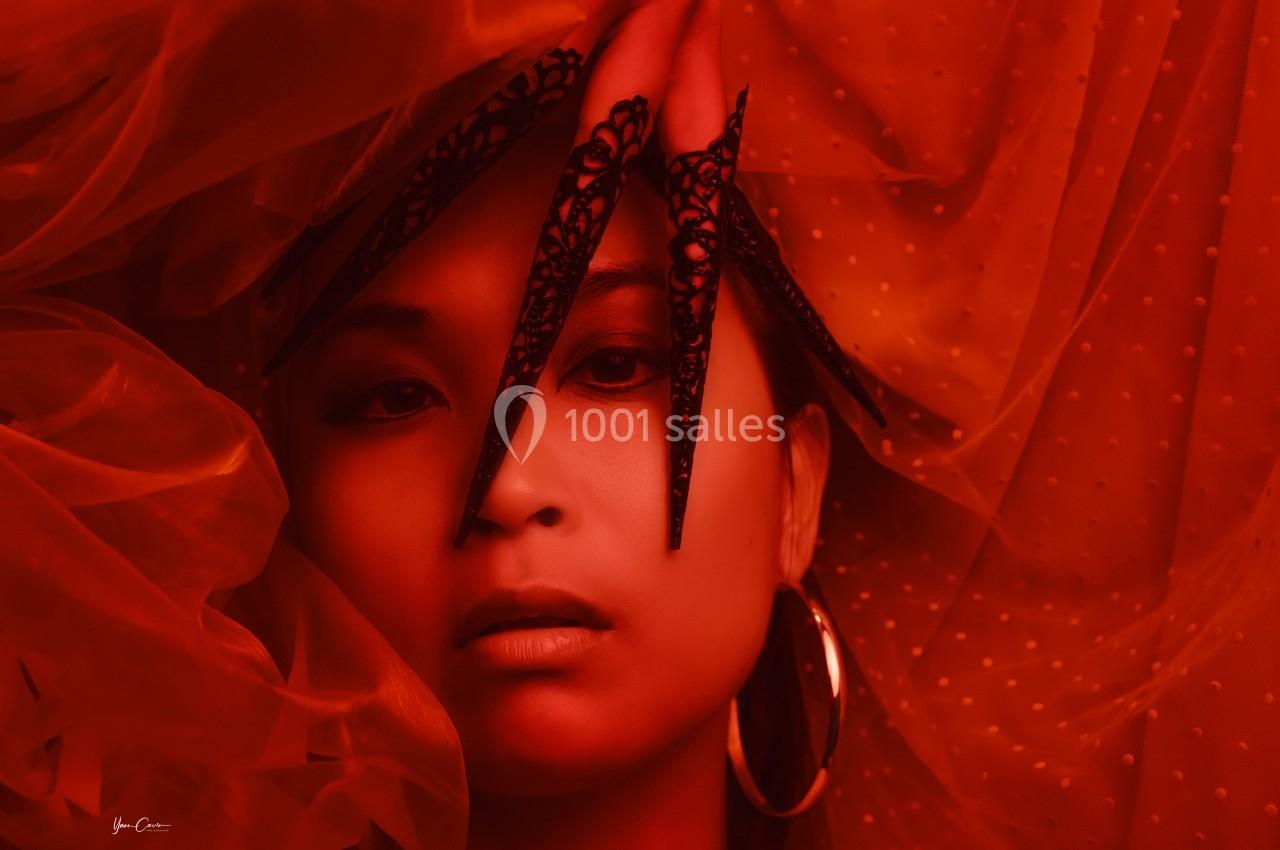 Femme portant des ornements noirs sur les doigts, éclairée par une lumière rouge intense avec un voile en arrière-plan.