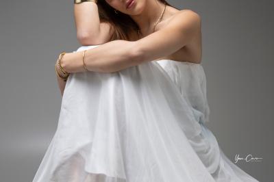 Miniature Shooting Portrait Femme assise, vêtue d'une robe blanche fluide, posant avec une expression sereine sur fond neutre.