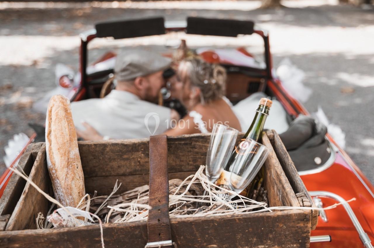 Un couple assis dans une voiture ancienne, avec une caisse en bois contenant une baguette, des flûtes et une bouteille.