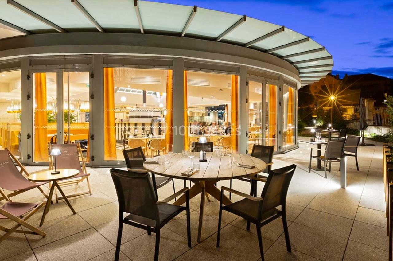 Terrasse d'un restaurant moderne avec tables et chaises, éclairée en soirée, vue sur l'intérieur lumineux à travers de…