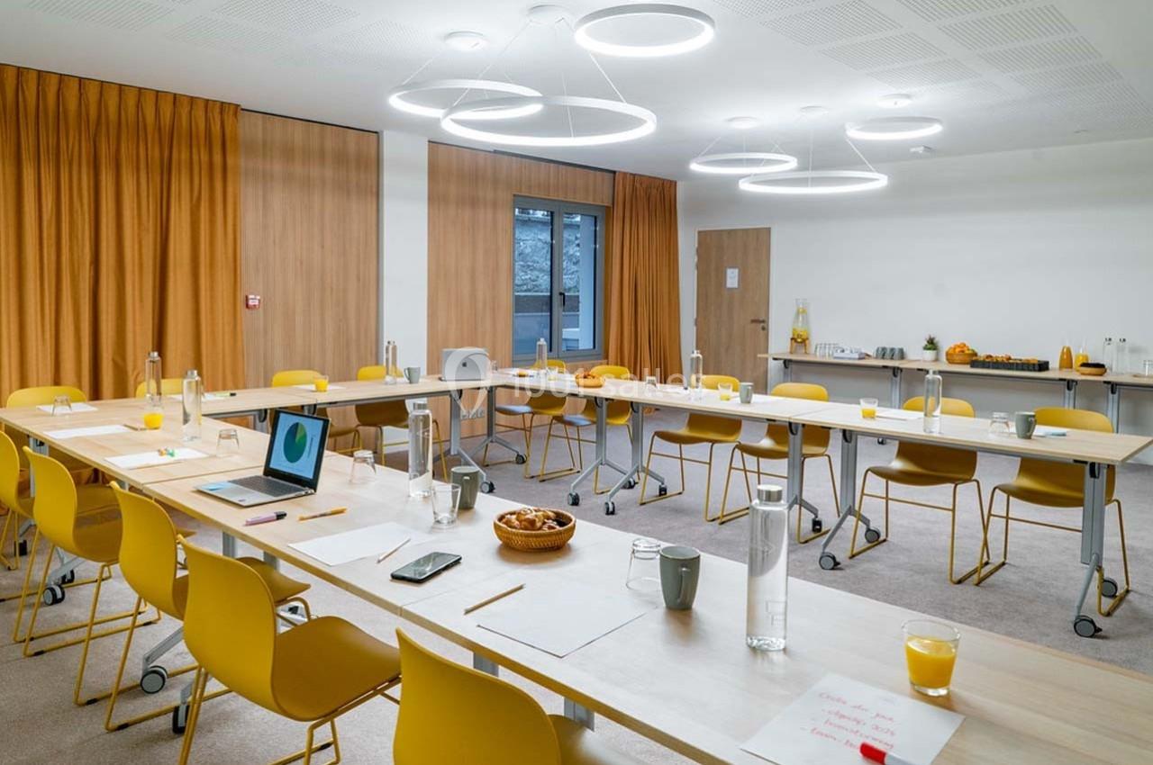 Salle de réunion lumineuse avec tables disposées en U, chaises jaunes, matériel de travail et buffet en arrière-plan.