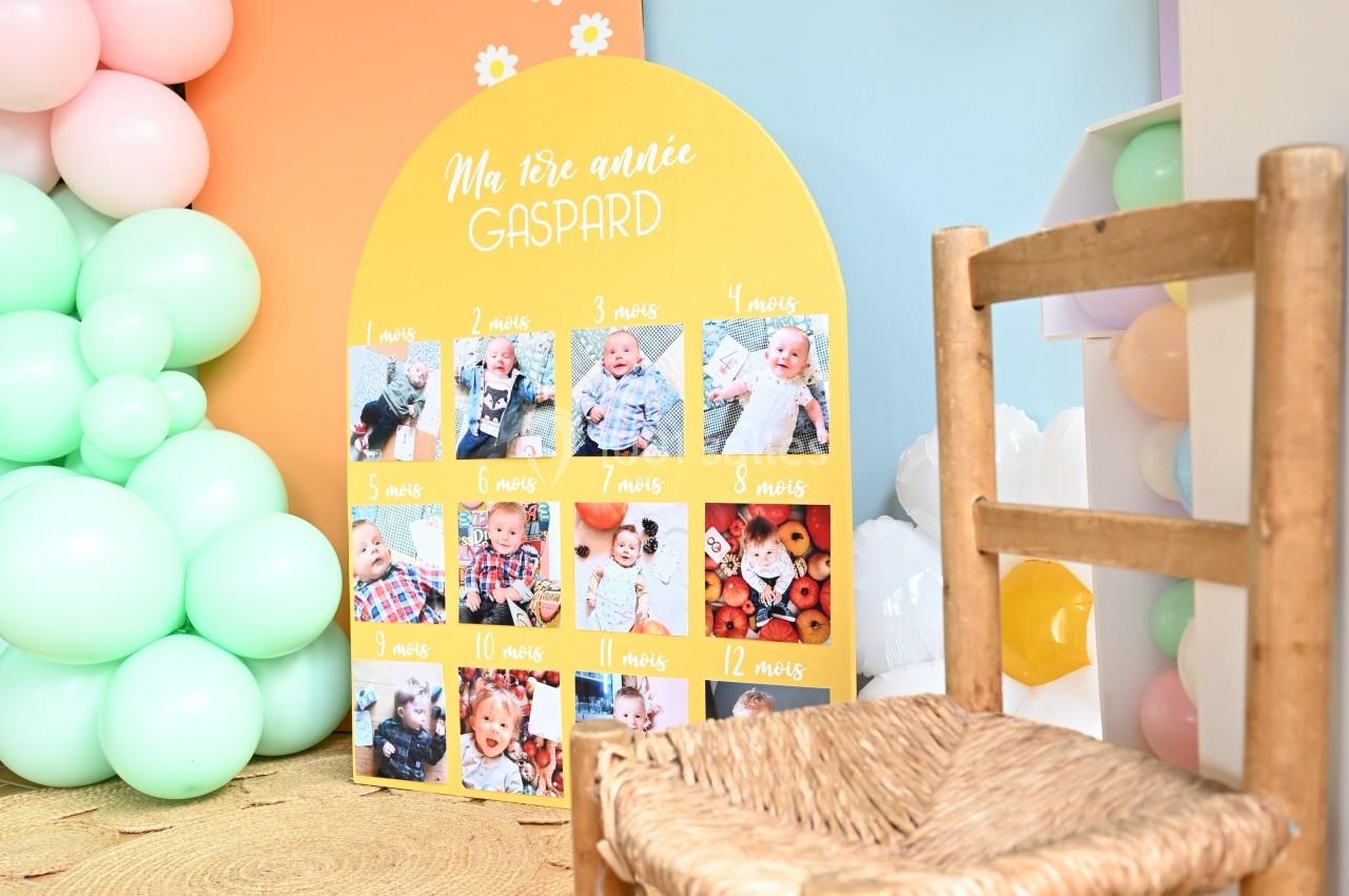 Panneau coloré illustrant les étapes de la première année d'un enfant, entouré de ballons pastel et d'une chaise en osier.