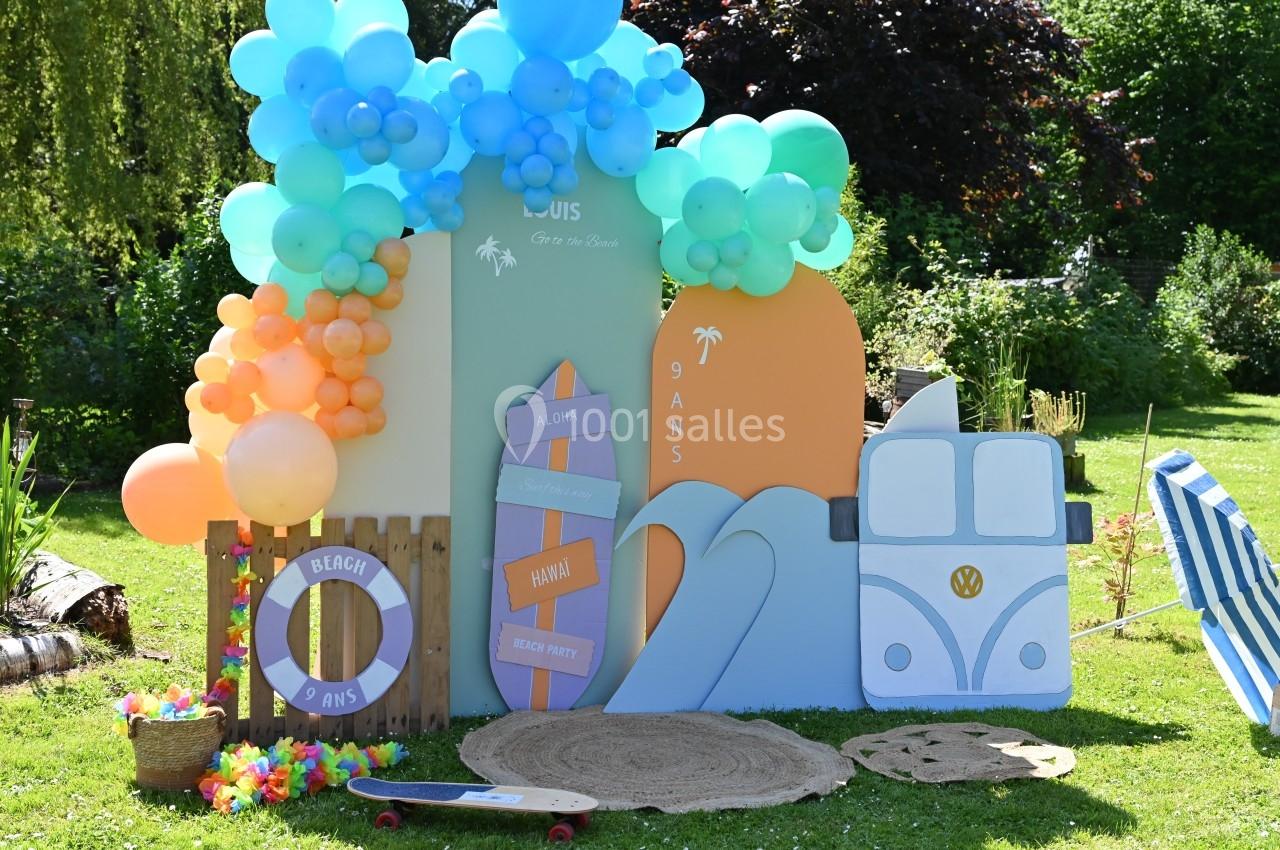 Décor festif sur le thème de la plage avec ballons colorés, planche de surf, van et accessoires estivaux dans un jardin.