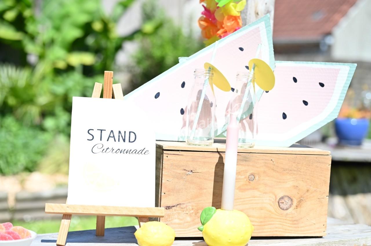 Stand de limonade avec pancarte sur chevalet, bouteilles décorées et accessoires en forme de citron et pastèque.