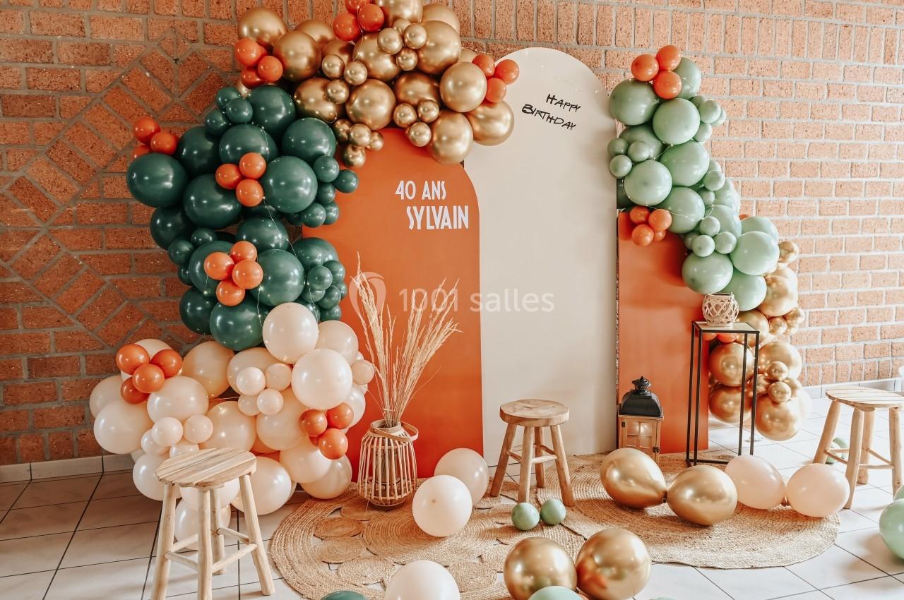 Décoration d'anniversaire avec ballons verts, oranges et dorés, panneaux personnalisés et tabourets en bois.