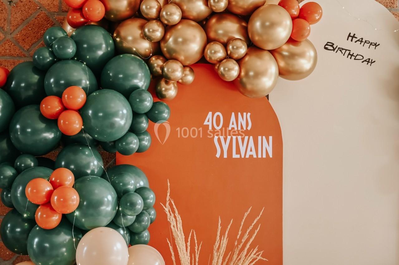 Décoration d'anniversaire avec ballons verts, orange et dorés, panneau ’40 ans Sylvain’ et inscription ’Happy Birthday’.