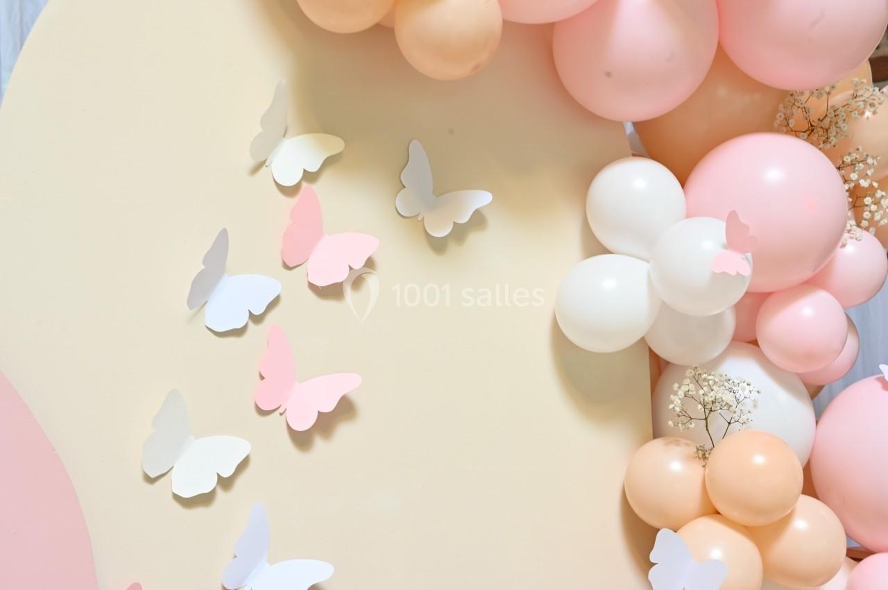 Décoration avec ballons pastel et papillons en papier sur fond beige, évoquant une ambiance douce et festive.