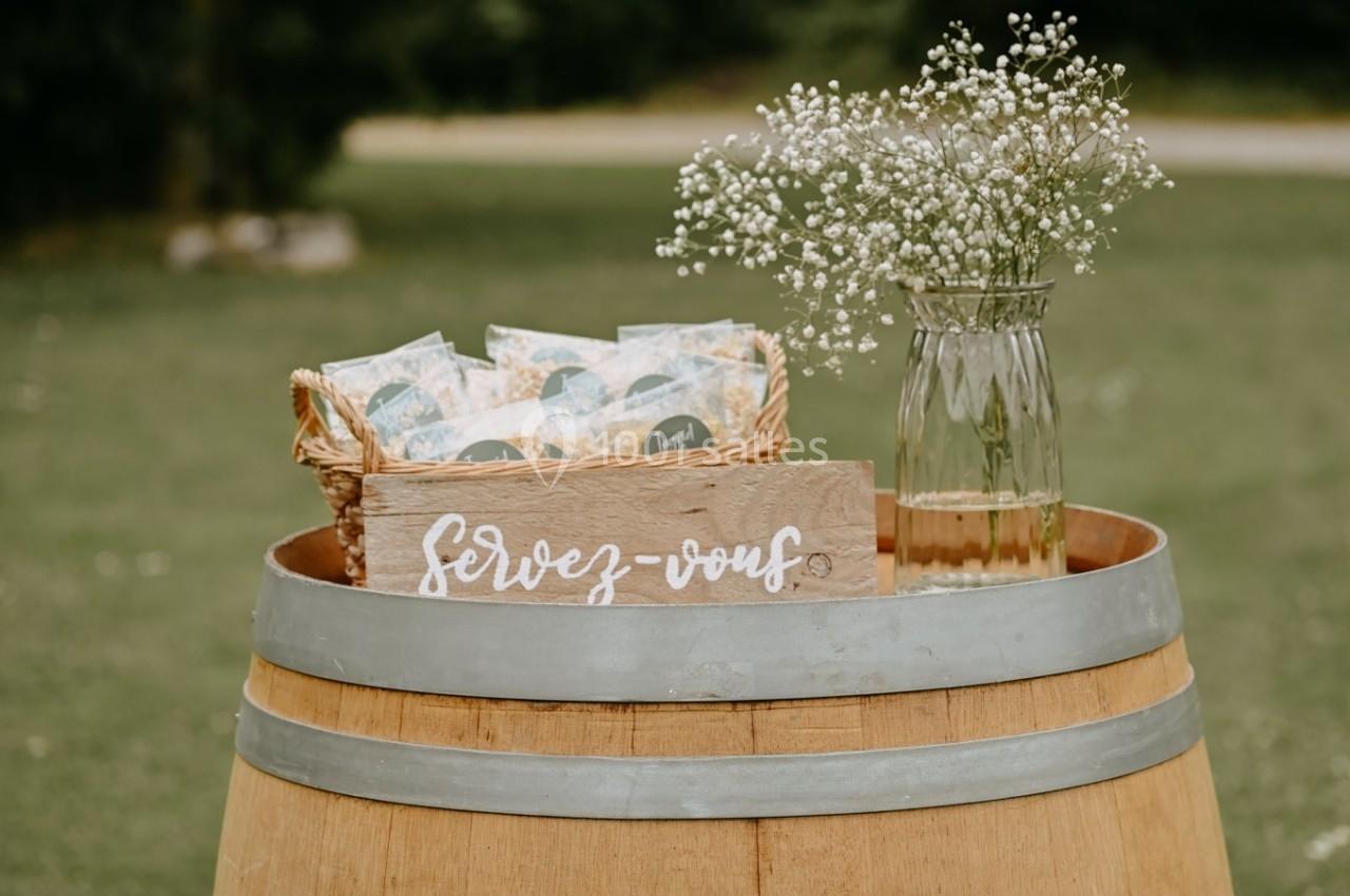 Panier en osier rempli de sachets, posé sur un tonneau avec une pancarte ’Servez-vous’ et un vase de fleurs blanches.