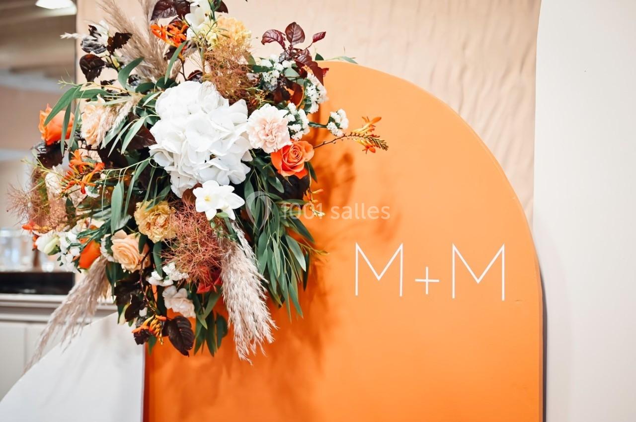 Arrangement floral coloré avec roses, feuillages et fleurs blanches, placé devant un panneau orange portant l'inscription…