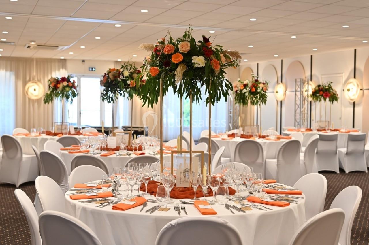 Salle de réception élégamment décorée avec des tables rondes, nappes blanches, centres de table fleuris et serviettes orange.