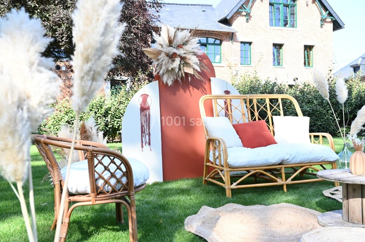 Salon de jardin en rotin avec coussins blancs et orange, décoré de pampas, installé sur une pelouse devant une maison en…